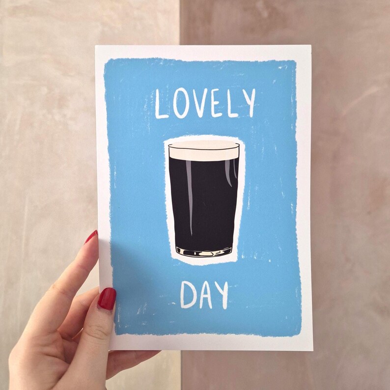 May include: A rectangular greeting card with a light blue background featuring a centered illustration of a dark stout beer in a glass with a white head. The text LOVELY is printed in white above the glass and the word DAY is printed below it.