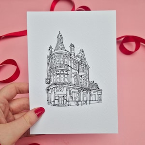 May include: A black and white line drawing of a building with the text "KINGS HEAD" on a white card. The card is held by a hand with red nail polish. Red ribbon is in the background.