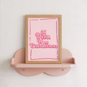 May include: A light pink rectangular print in a light wood frame featuring the dark pink text A Diva Has Distractions in a retro font style. The print includes a hand-drawn dark pink border and small white star accents on a light pink background.
