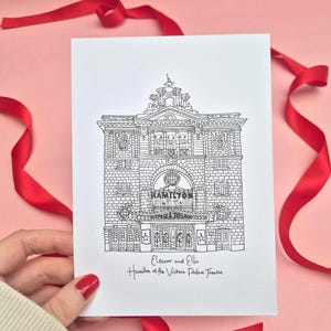 May include: A black and white line drawing of the Victoria Palace Theatre, with the word "HAMILTON" above the entrance. The card is held against a pink background with red ribbon. The text "Eleanor and Ellis" and "Hamilton at the Victoria Palace Theatre" is also visible.