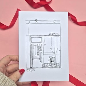 May include: A black and white line drawing of a building facade with an open door, two chairs, and hanging lights. The drawing is on white paper, held by a hand with red nail polish, against a pink background with red ribbon.