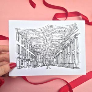 May include: Black and white line drawing of a street scene with buildings and string lights. The card is held against a pink background with red ribbon. The illustration depicts a detailed architectural view.