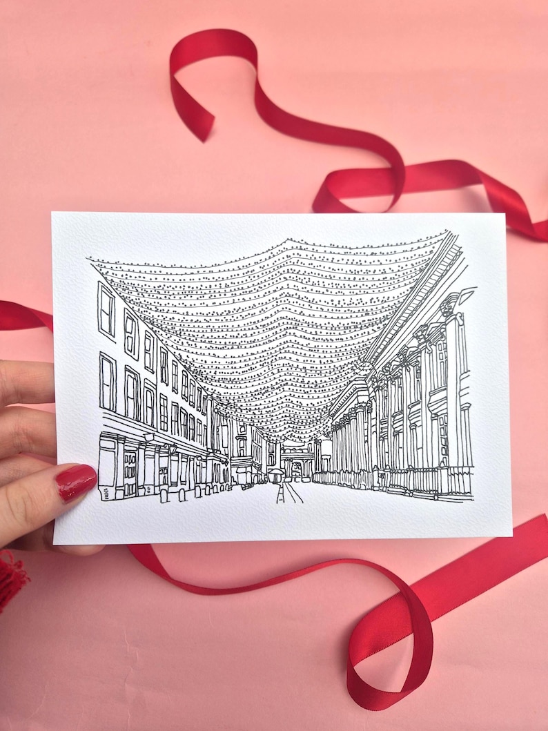 May include: Black and white line drawing of a street scene with buildings and string lights. The card is held against a pink background with red ribbon. The illustration depicts a detailed architectural view.