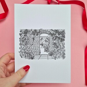 May include: A black and white line drawing of a garden scene on a white card. The illustration depicts a brick wall with an arched doorway, surrounded by flowers and foliage. A red ribbon is in the background.