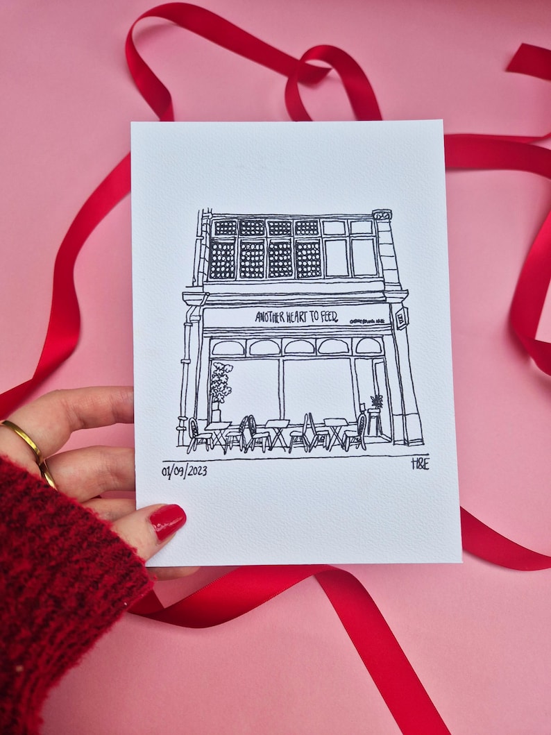 May include: A black and white line drawing of a cafe with the text "ANOTHER HEART TO FEED". The drawing includes tables, chairs, and a date. The artwork is on white paper, with a pink background and red ribbon.
