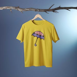 May include: A yellow t-shirt featuring a cartoon umbrella design. The umbrella has a pink top, purple trim, and yellow flower accents. The handle is purple. The shirt is hanging on a wooden hanger.