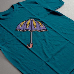 May include: Teal t-shirt featuring a cartoon umbrella design. The umbrella has a purple canopy with yellow and oval accents, a gray handle, and a pink curved handle. The design is centered on the shirt.