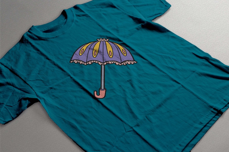 May include: Teal t-shirt featuring a cartoon umbrella design. The umbrella has a purple canopy with yellow and oval accents, a gray handle, and a pink curved handle. The design is centered on the shirt.