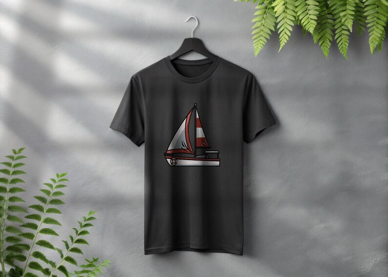 Peut inclure: T-shirt noir avec un motif de voilier. Le voilier a une voile blanche avec des rayures rouges et des bordures rouges. Le t-shirt est suspendu &agrave; un cintre noir sur un mur gris avec des plantes vertes.
