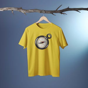 Peut inclure: Un t-shirt jaune avec un motif de boussole. La boussole est noire et argent&eacute;e avec une aiguille rouge. Le t-shirt est suspendu &agrave; un cintre en bois sur un fond bleu.
