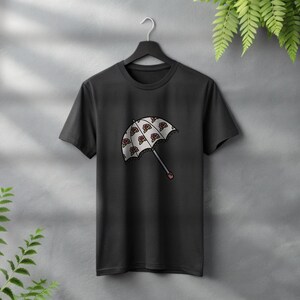 May include: Black t-shirt featuring a graphic of a white umbrella with a repeating pattern of red and brown shapes. The umbrella has a gray handle with a red tip. The shirt is hanging on a black hanger.