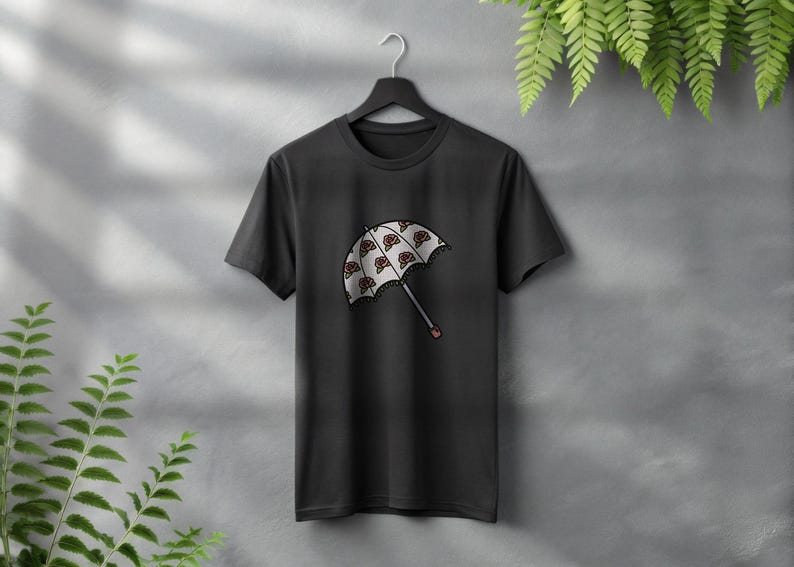 May include: Black t-shirt featuring a graphic of a white umbrella with a repeating pattern of red and brown shapes. The umbrella has a gray handle with a red tip. The shirt is hanging on a black hanger.