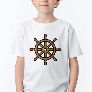 Peut inclure: Un t-shirt blanc avec un motif de gouvernail de navire marron et dor&eacute;. Le motif est centr&eacute; sur le t-shirt. La personne qui porte le t-shirt a les cheveux roux et sourit.
