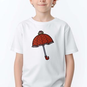May include: White t-shirt featuring a cartoon umbrella design. The umbrella has a red polka-dot canopy, a silver handle, and a red curved handle. The shirt is worn by a child with red hair.