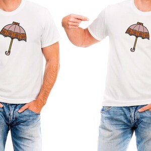 May include: White t-shirt featuring a cartoon umbrella design. The umbrella has a pink canopy with a decorative border, a yellow handle, and a curved handle end. The shirt is paired with blue jeans.