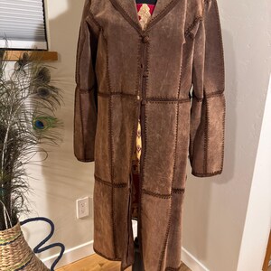 May include: A long, brown suede coat with decorative stitching. The coat has a notched collar, button closures, and paneled construction. The coat is displayed on a black mannequin stand.