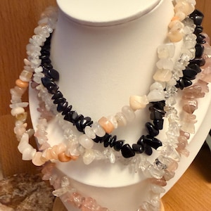 May include: A selection of gemstone chip necklaces in shades of clear, black, peach, and pink. The necklaces are displayed on a white bust, highlighting the different stone colours and textures. These necklaces are a unique jewellery item.