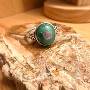 May include: A silver ring featuring a large, round, green gemstone with a pinkish-red centre. The ring has decorative filigree on the sides. The ring is displayed on a light brown, textured surface.