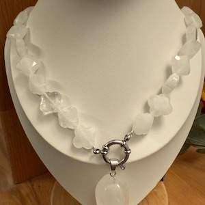 May include: A white quartz necklace displayed on a white mannequin. The necklace features irregularly shaped quartz beads and a large, teardrop-shaped pendant. A silver-colored clasp secures the necklace.