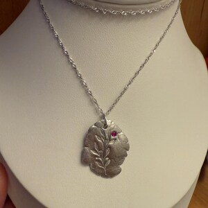 Op de afbeelding: Een zilverkleurige ketting met een blad-vormige hanger. De hanger heeft een getextureerd oppervlak en een kleine, ronde, rode edelsteen. De ketting wordt getoond op een witte mannequin.