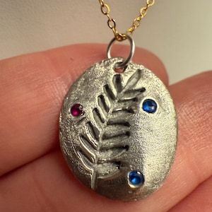 Può includere: Collana con pendente argentato e catena dorata. Il pendente ovale ha una superficie strutturata, un motivo a foglie e tre pietre preziose colorate: una rossa e due blu. La collana è un gioiello.
