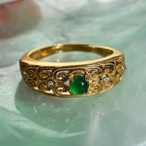 Könnte beinhalten: Ein goldfarbener Ring mit einem durchbrochenen Filigranband mit Wirbelmustern. In der Mitte befindet sich ein kleiner, runder, leuchtend grüner Cabochon-Stein, flankiert von zwei winzigen, klaren, funkelnden Akzentsteinen im Metall.
