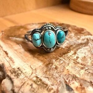 Puede incluir: Un anillo de plata con tres piedras turquesas ovaladas. Las piedras están engastadas en un engaste decorativo y ornamentado. El anillo se exhibe sobre una superficie texturizada de color marrón claro.