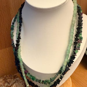 Könnte beinhalten: Drei Halsketten auf einer weißen Büste. Die Halsketten bestehen aus sternförmigen Perlen in Grün-, Schwarz- und Rottönen. Die Halsketten werden auf einer weißen Büste vor einem Holzgrund präsentiert.