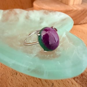 Op de afbeelding: Een zilveren ring met een grote, ovale edelsteen, diep paars in het midden en groen aan de randen. De ring ligt op een lichtgroen, hartvormig oppervlak, met een houten achtergrond.