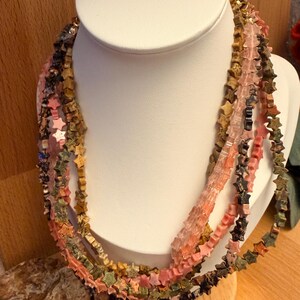 May include: A multi-strand necklace on a white display bust. The necklace has star-shaped beads in pink, green, brown, and black. The beads are strung together, creating a layered effect. A unique jewellery design.