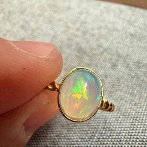 Könnte beinhalten: Ein goldener Ring mit einem ovalen Opal-Edelstein. Der Opal zeigt ein Farbspiel mit grünen, gelben und orangenen Blitzen. Der Ring hat ein gedrehtes Seil-Design.