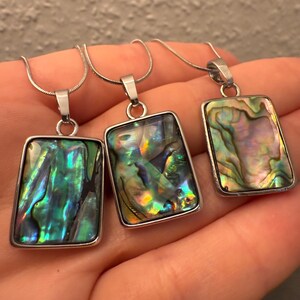 Könnte beinhalten: Drei Halsketten mit rechteckigen Anhängern aus schillerndem Abalone-Muschelmaterial, gefasst in silberfarbenen Rahmen. Die Abalone-Muscheln zeigen Farben wie Grün, Blau und Lila. Jeder Anhänger hängt an einer feinen Silberkette.