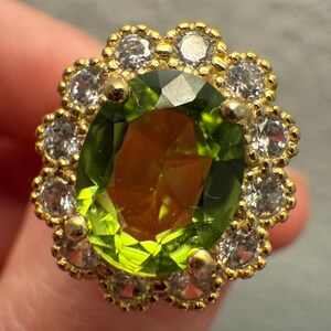 Könnte beinhalten: Ein goldfarbener Ring mit einem großen, oval geschliffenen, hellgrünen Edelstein im Zentrum. Der Edelstein ist von einem Kranz kleiner, klarer, rund geschliffener Steine umgeben. Die Fassung des Rings hat ein detailliertes, blumenähnliches Design.