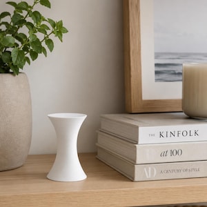 May include: A small, matte white candle holder with a smooth, hourglass-shaped silhouette. The object features a wide, circular base and a matching wide top, tapering inward toward the center, creating a clean, symmetrical, and solid form.
