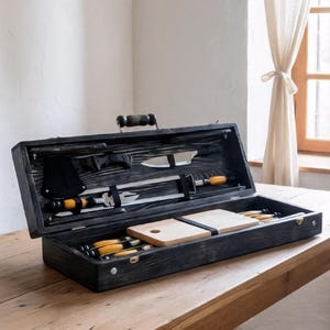 May include: A black wooden barbecue tool set, open on a wooden table. The set includes knives, a carving fork, and cutting boards. The tools have wooden handles and are secured inside the box. The box has a handle.
