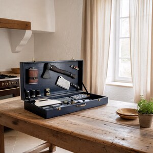May include: A black briefcase, open to reveal a collection of tools, including an axe and cleaver. The case sits on a wooden table. The scene is set in a kitchen, with a window and curtains visible in the background. The case is open.