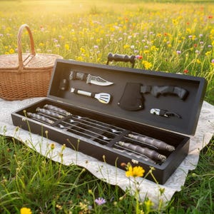 May include: A black wooden barbecue tool set containing skewers, a knife, a small axe, and a bottle opener. The set is open on a white cloth in a field of wildflowers, with a wicker basket in the background. The tools are stainless steel.