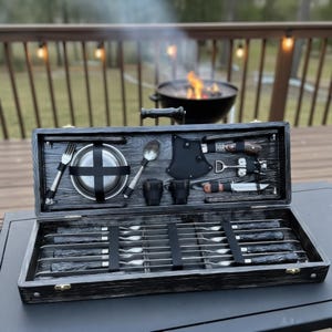 May include: A black wooden barbecue utensil set with a handle, fork, spoon, bowl, cups, and knives. The set includes a small axe and is displayed open on a table. A grill with a fire is in the background.