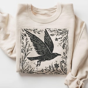 Könnte beinhalten: Cremefarbenes Sweatshirt mit einem schwarzen Grafikmotiv eines fliegenden Vogels, umrahmt von botanischen Elementen. Das Design befindet sich mittig auf der Vorderseite des gefalteten Kleidungsstücks. Die Ärmel sind hochgekrempelt, der Stoff wirkt weich.