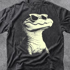 May include: Dark grey t-shirt with a graphic of an alligator wearing sunglasses. The alligator is in a light colour, contrasting with the shirt's dark background. The word "PEPPER" is at the top left. The brand "OLD PINES CO" is at the bottom right.
