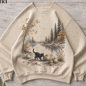 Pode incluir: Uma camisola de gola redonda cor de areia com uma impressão gráfica de um gato preto perto de abóboras junto a um lago. A cena inclui uma floresta com pinheiros, folhagem de outono e pássaros no céu. A palavra Sand está no canto superior.