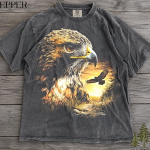 Może przedstawiać: Ciemnoszary t-shirt z krótkim rękawem z dużym nadrukiem graficznym przedstawiającym głowę jastrzębia z profilu, ze złocistobrązowymi piórami i żółtym dziobem. Mniejszy jastrząb leci na tle zachodzącego słońca z wysoką trawą na pierwszym planie.