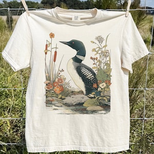 May include: Off-white t-shirt with a detailed illustration of a loon bird amongst flowers and reeds. The bird has a black head and back with a white and black speckled body. The shirt is hanging on a clothesline.