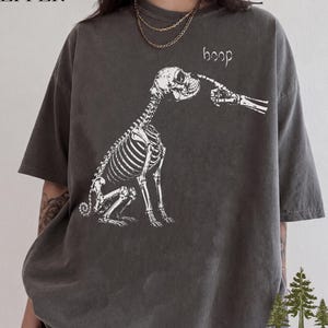Könnte beinhalten: Ein anthrazitfarbenes Kurzarm-T-Shirt mit einer weißen Grafik eines Hundeskeletts, das mit einem skelettierten Finger an der Nase berührt wird. Der Text "boop" ist in einer handschriftlichen Schriftart über dem Kopf des Hundes aufgedruckt.