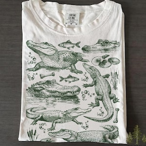 Puede incluir: Camiseta blanca con una ilustración verde de caimanes, peces y plantas acuáticas. El diseño incluye varias poses de caimanes y detalles como burbujas y huesos. La camiseta tiene cuello redondo y mangas cortas. La etiqueta dice "Comfort Colors".