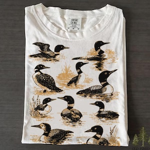 May include: White crew neck t-shirt with a detailed illustration of loons in a water scene. The design is in black and tan. The brand "Comfort Colors" is visible at the top. The t-shirt is folded.