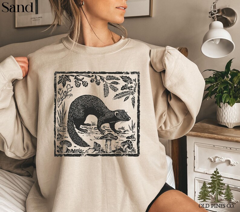 May include: Sand-colored sweatshirt featuring a black and white illustration of a ferret surrounded by plants and mushrooms within a rectangular frame. The text "Sand" is in the upper left corner. The sweatshirt is from Old Pines Co.