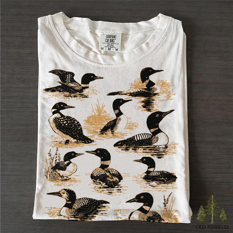 May include: White t-shirt with a crew neck, featuring a detailed illustration of various loons in a water scene. The design is in black and tan, with the brand "Comfort Colors" visible at the top. The shirt is folded.