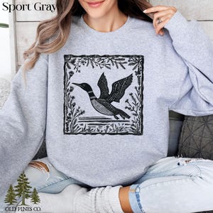 May include: Sport Gray sweatshirt featuring a black and white illustration of a loon in flight, framed by a floral border. The sweatshirt has the text "Sport Gray" at the top and the "OLD PINES CO." logo at the bottom.