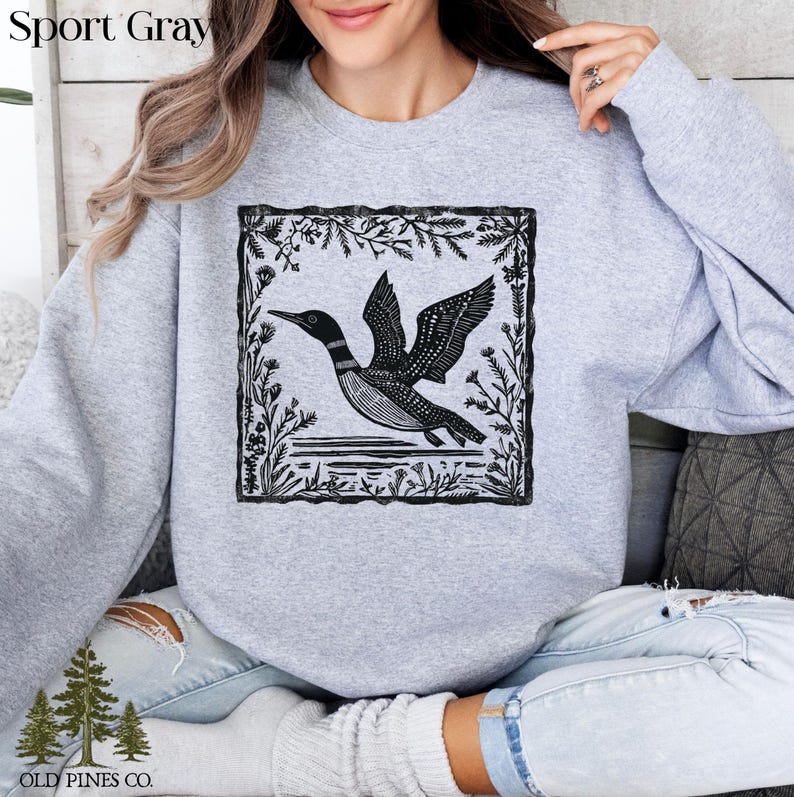 May include: Sport Gray sweatshirt featuring a black and white illustration of a loon in flight, framed by a floral border. The sweatshirt has the text "Sport Gray" at the top and the "OLD PINES CO." logo at the bottom.